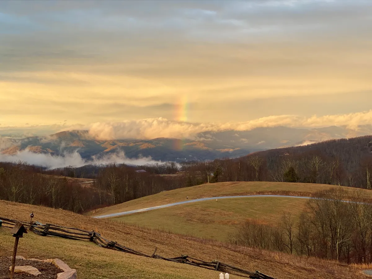 A sweeping mountain view unfolds beyond a rustic wooden fence, with a rainbow arching over rolling hills and dramatic clouds. The serene atmosphere is ideal for reflection, photography, or picnics during festival days.