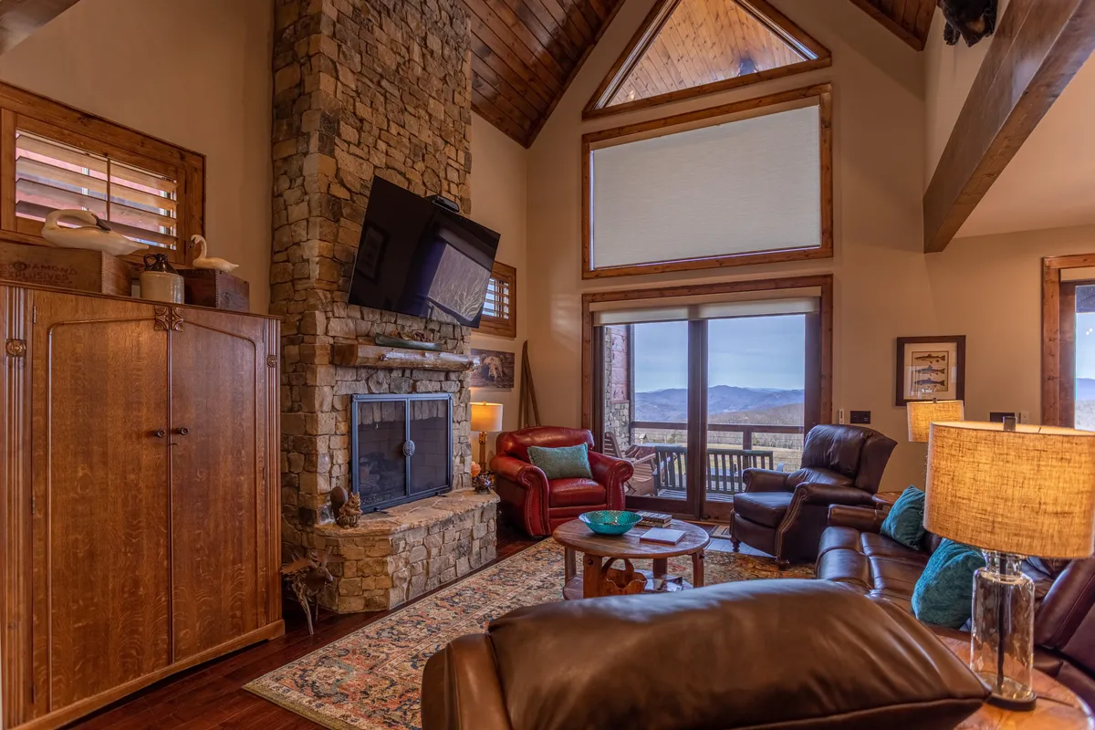 A cozy living area with a stone fireplace, comfortable seating, large windows that frame stunning mountain views, and a decorative table. The mood is warm and welcoming, suggesting evenings of relaxation and togetherness by the fire, or enjoying the scenery.