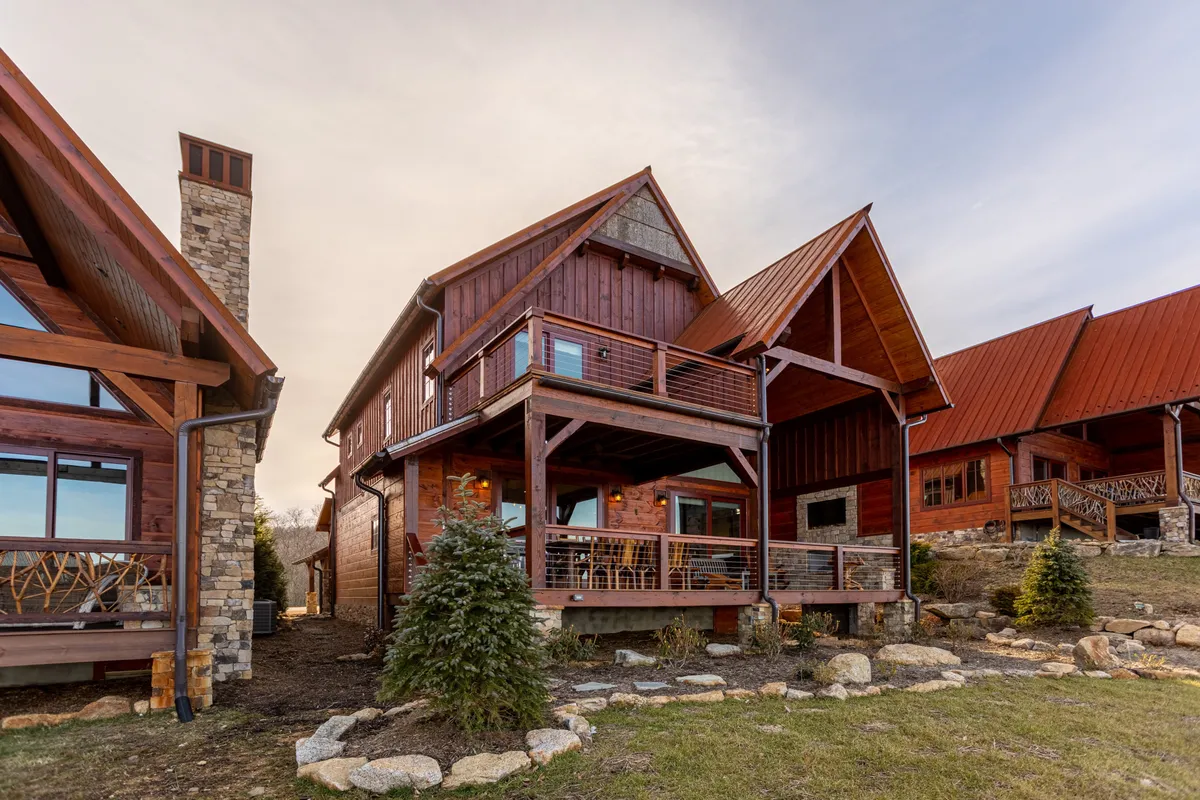 This exterior image highlights the wooden balcony of Whitetail Ridge Lodge, with a stone chimney, large windows, and a landscaped yard set against a backdrop of natural beauty. The vantage point suggests a perfect spot for watching sunsets, fireworks, or gathering with family for celebrations.
