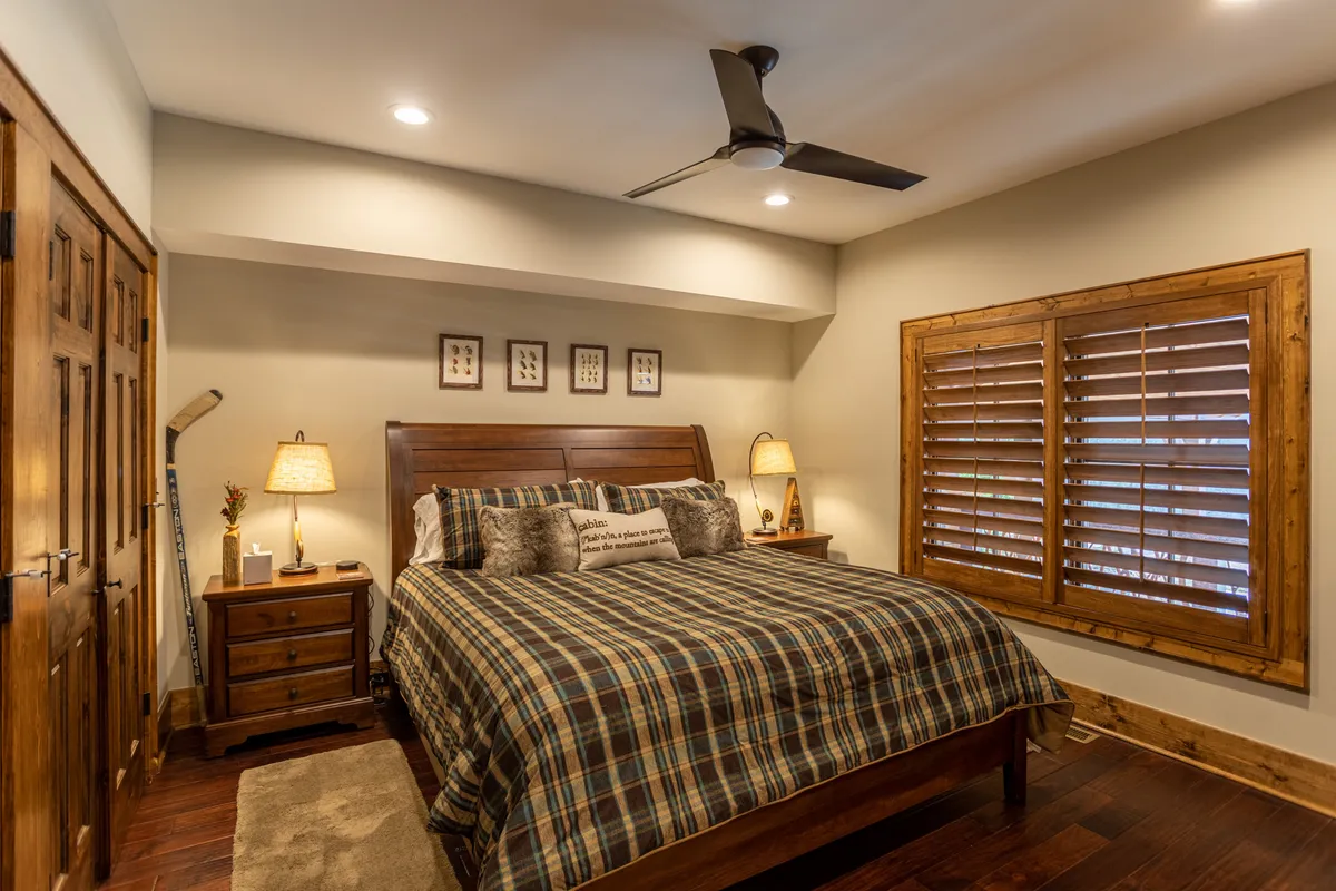 A comfortable bedroom showcasing a wooden bed frame with plaid bedding, decorative pillows, a nightstand with a lamp, and a shuttered window letting in gentle natural light. The intimate setting exudes warmth and relaxation, ideal for a peaceful night's sleep.
