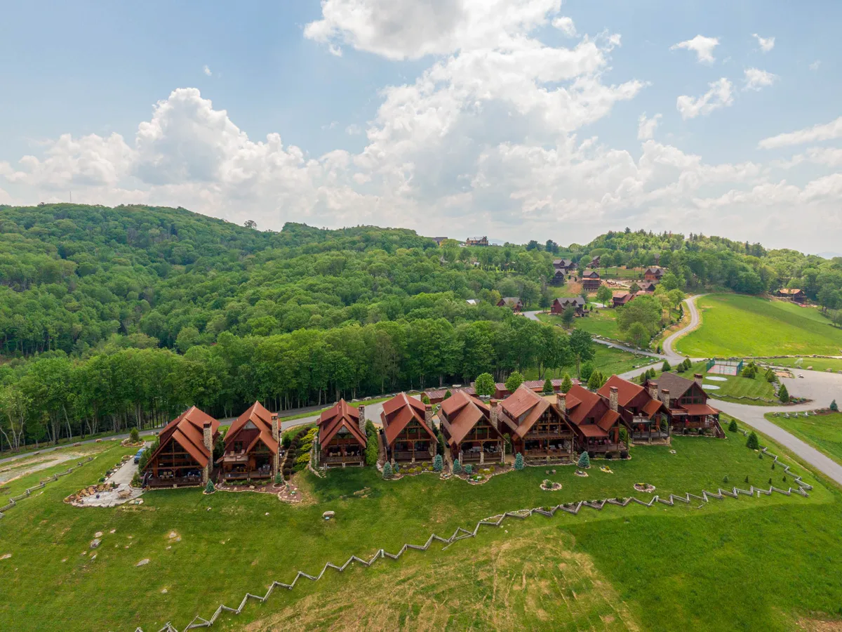 This image captures the charming exterior of wooden chalets nestled in a lush green landscape, with a winding road leading towards distant hills under an open sky. The tranquil scene suggests both seclusion and access to resort amenities, with space for outdoor gatherings or a romantic stroll under the stars.