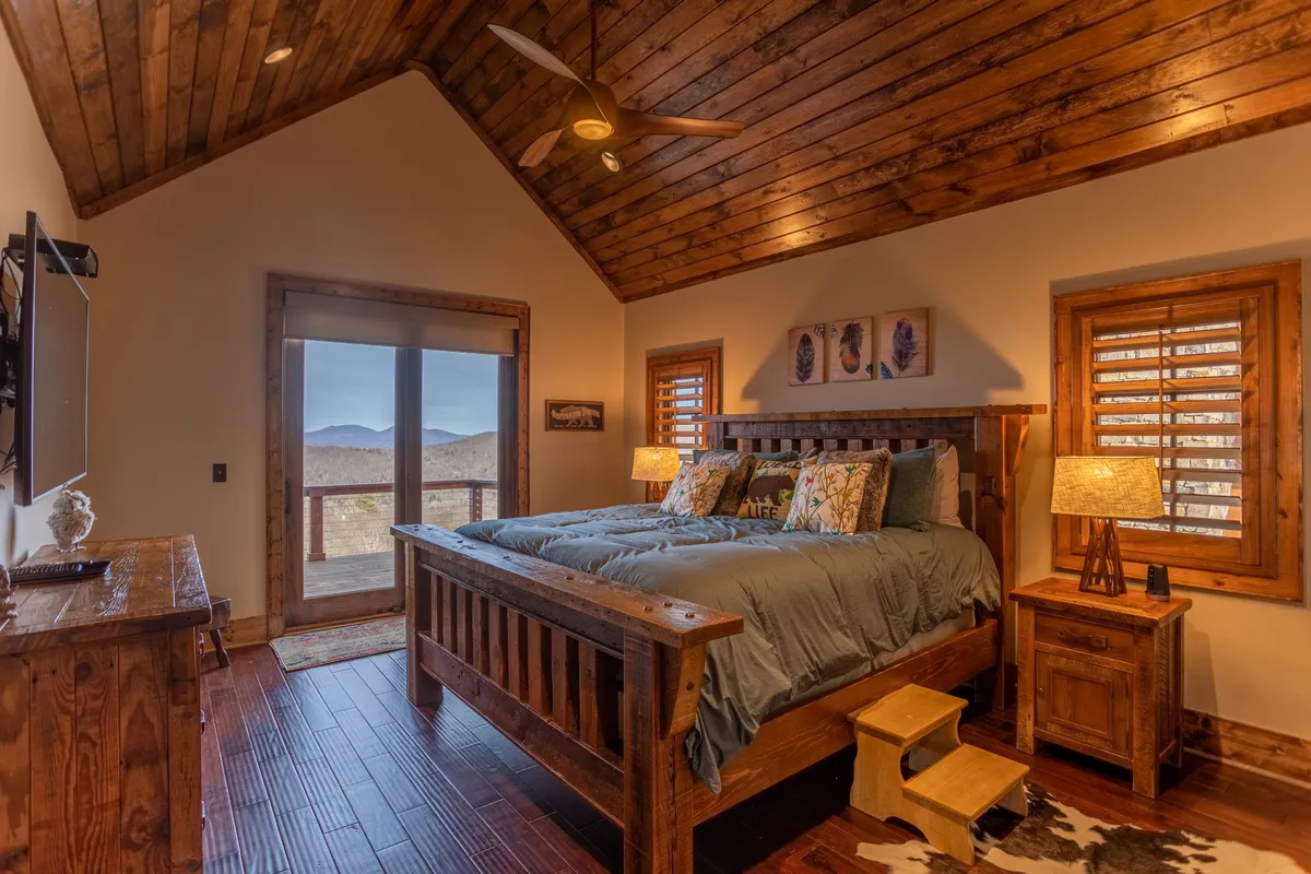 A spacious king suite featuring a king-sized bed adorned with decorative pillows, large windows that frame stunning mountain views, and rustic wooden furniture. The serene atmosphere invites relaxation, with natural light streaming in and a cozy reading nook by the window.
