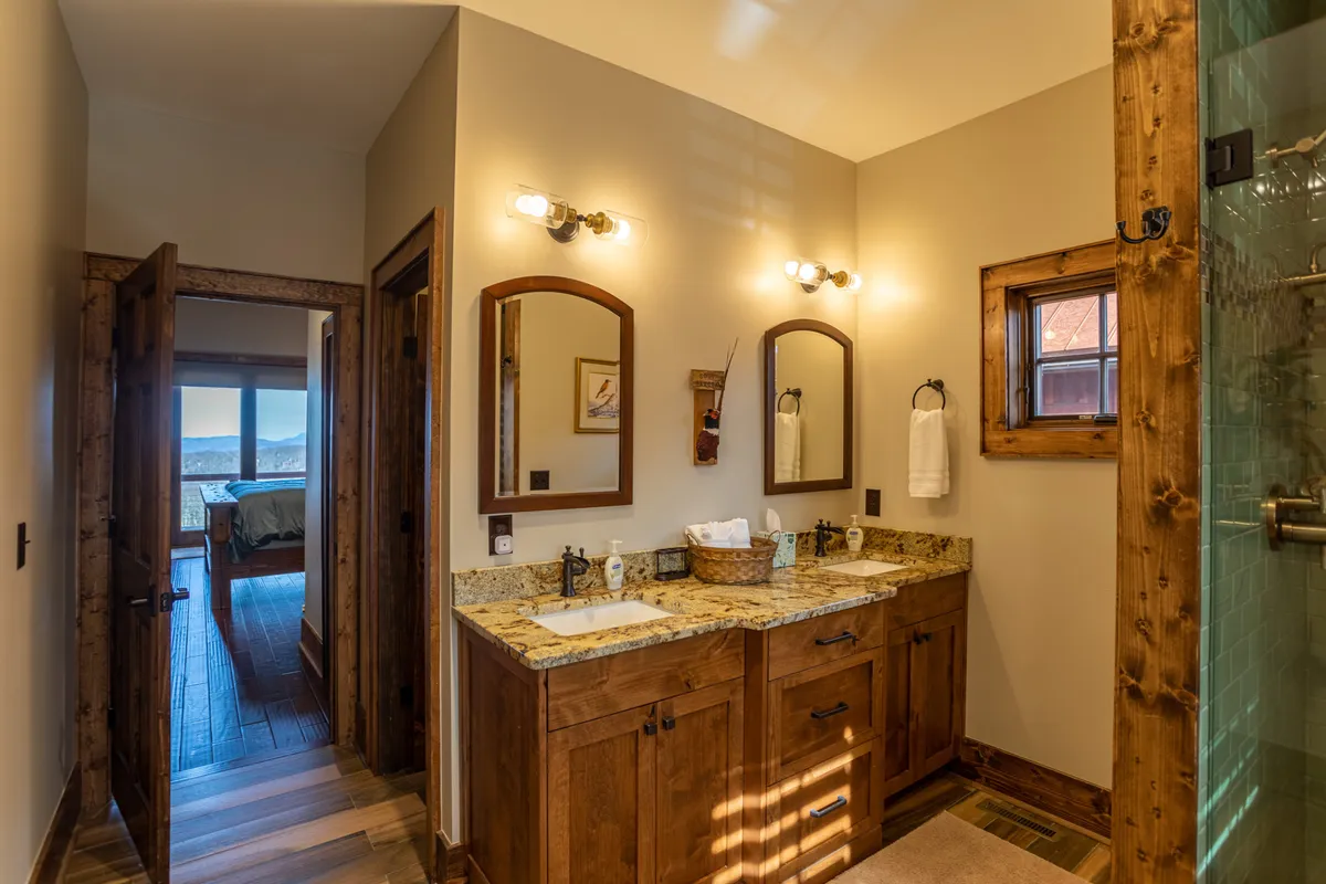 A welcoming bathroom with a wooden vanity, granite countertop, framed mirrors, and a spacious shower area. A window offers natural light and a view to the outdoors, enhancing the sense of calm and privacy.