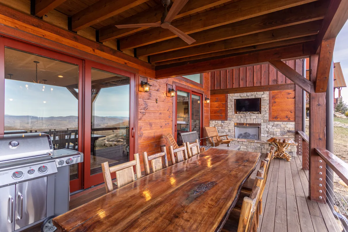 This photo features a large wooden dining table set for outdoor meals, a grill for barbecuing, a stone outdoor fireplace, cushioned seating, and large windows looking back into the home. The covered structure enhances its use from sunny mornings to crisp evenings, and the mountain backdrop adds to the ambiance.