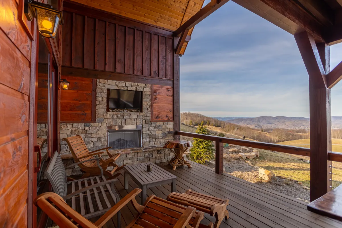 This image showcases a spacious wooden deck outfitted with comfortable outdoor seating, a prominent stone fireplace, a mounted outdoor television, and sweeping views of the mountains beyond. The deck is partially covered, making it suitable for all-weather enjoyment and social gatherings.