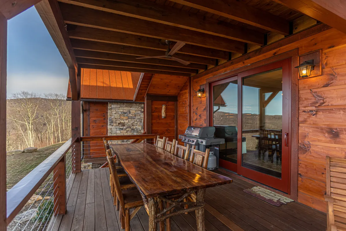 An inviting exterior shot of a deck set with a large wooden table for group dining, an outdoor grill for barbecues, and a scenic backdrop of rolling hills and mountains. Wooden beams frame the deck, enhancing its rustic charm, while the setup suggests lively gatherings and relaxation.