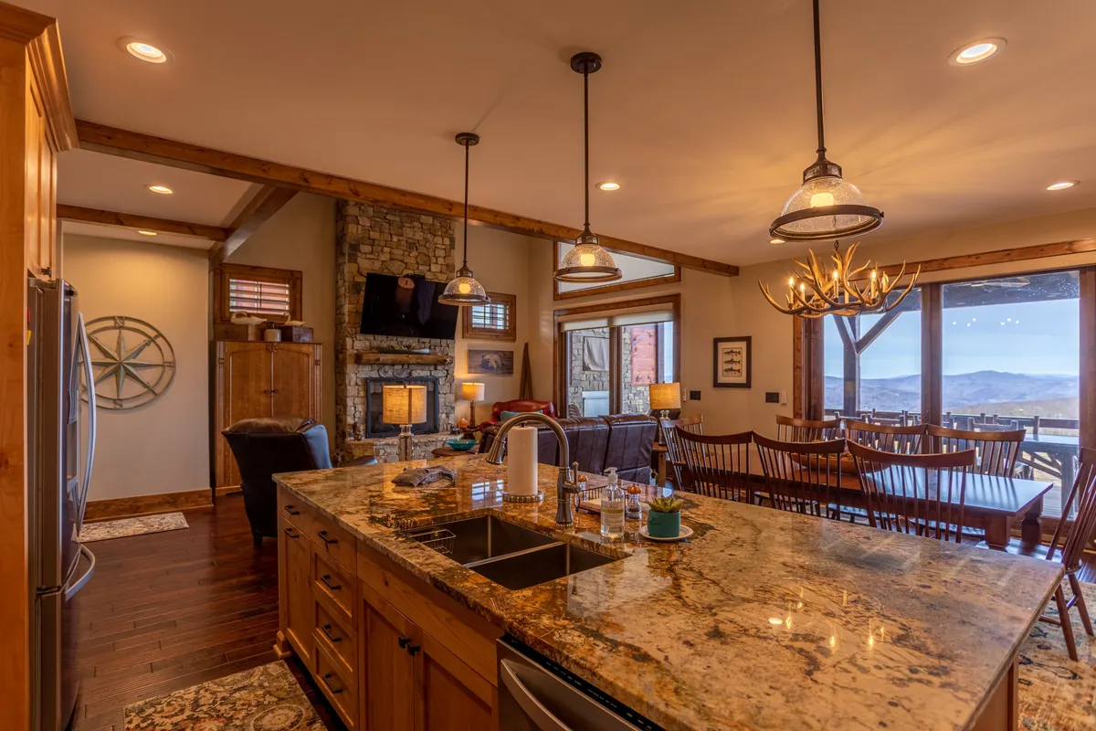 This image presents a seamless view of the kitchen, dining area, and living room. The granite countertop is visible along with a stone fireplace, large windows offering a mountain view, a spacious dining area, and a beautiful chandelier. The layout showcases the openness and togetherness described in the section, with visual cues for family gatherings and entertaining.