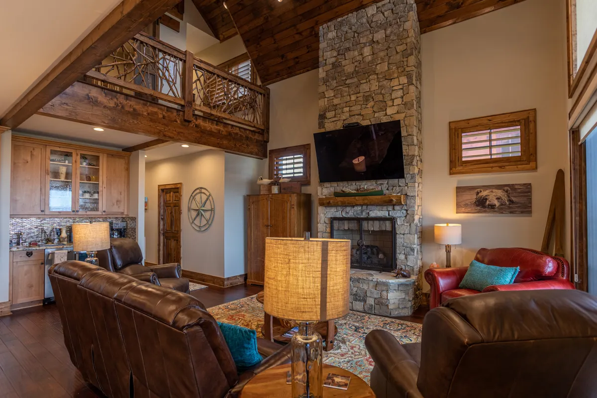 This image showcases a spacious living area featuring a stone fireplace, plush leather armchairs, wooden beams overhead, and large windows that likely frame scenic views beyond. The setting is ideal for gathering with friends after a day of outdoor adventure, relaxing by the fire, watching movies, or enjoying a warm drink together.